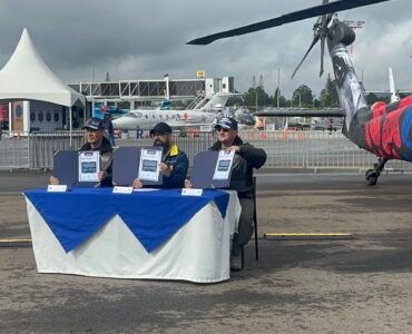 El MG ANDRÉS GUZMÁN JUNTO A OTRAS DOS PERSONAS MOSTRANDO EL RECONOCIMIENTO QUE LES ENTREGARON EN LA FERIA F-AIR 2025, SENTADOS EN UNA MESA AL AIRE LIBRE, AL FONDO VRIAS AERONAVES EN EXHIBICIÓN
