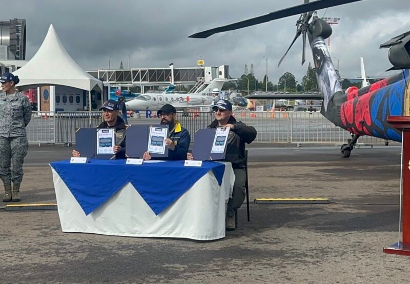 IMG_3028 El MG ANDRÉS GUZMÁN JUNTO A OTRAS DOS PERSONAS MOSTRANDO EL RECONOCIMIENTO QUE LES ENTREGARON EN LA FERIA F-AIR 2025, SENTADOS EN UNA MESA AL AIRE LIBRE, AL FONDO VRIAS AERONAVES EN EXHIBICIÓN