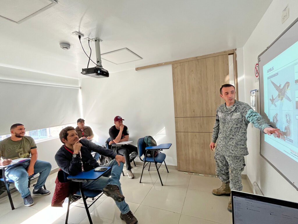 Un instructor de la Fuerza aérea perteneciente al equipo de mantenimiento de CIAC explicando sobre un curso de la aeronave C295 en un aula del centro de entrenamiento donde se ven 5 personas sentados prestando atención a su explicación, Desde una perspectiva lateral.