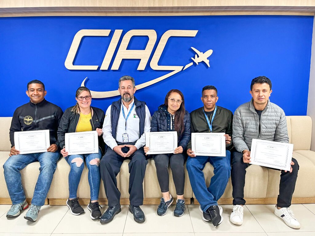 Cinco personas sosteniendo un diploma y en el centro el instructor del curso del cual recibieron el diploma, todos están sentados frente a una pared azul con el logo de CIAC.
